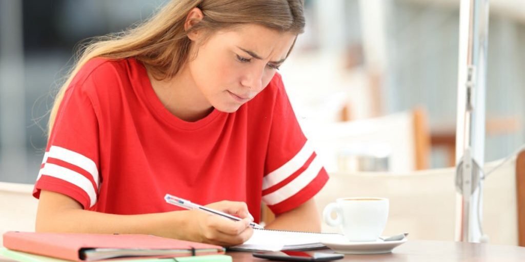 A girl is writing in the book and there is a cup next to her.