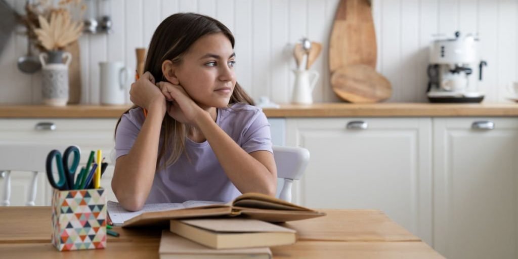 A girl is reading books and there are other books on the table, scissors and pencils.